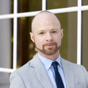 A light-skinned man in a light blue button down shirt crosses his arms and smiles.