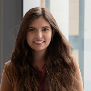 An image of Keisha Blain weaing a textured white jacket and a blue-green shirt. She wears twists just past her shoulders and black glasses as she smiles for the camera.