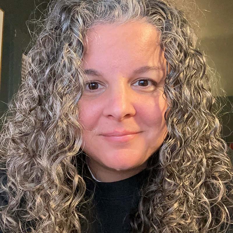 An image of Keisha Blain weaing a textured white jacket and a blue-green shirt. She wears twists just past her shoulders and black glasses as she smiles for the camera.