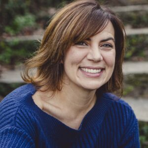 Liz Neeley, wearing a royal blue sweater and shoulder-length brown hair, smiles widley for the camera.