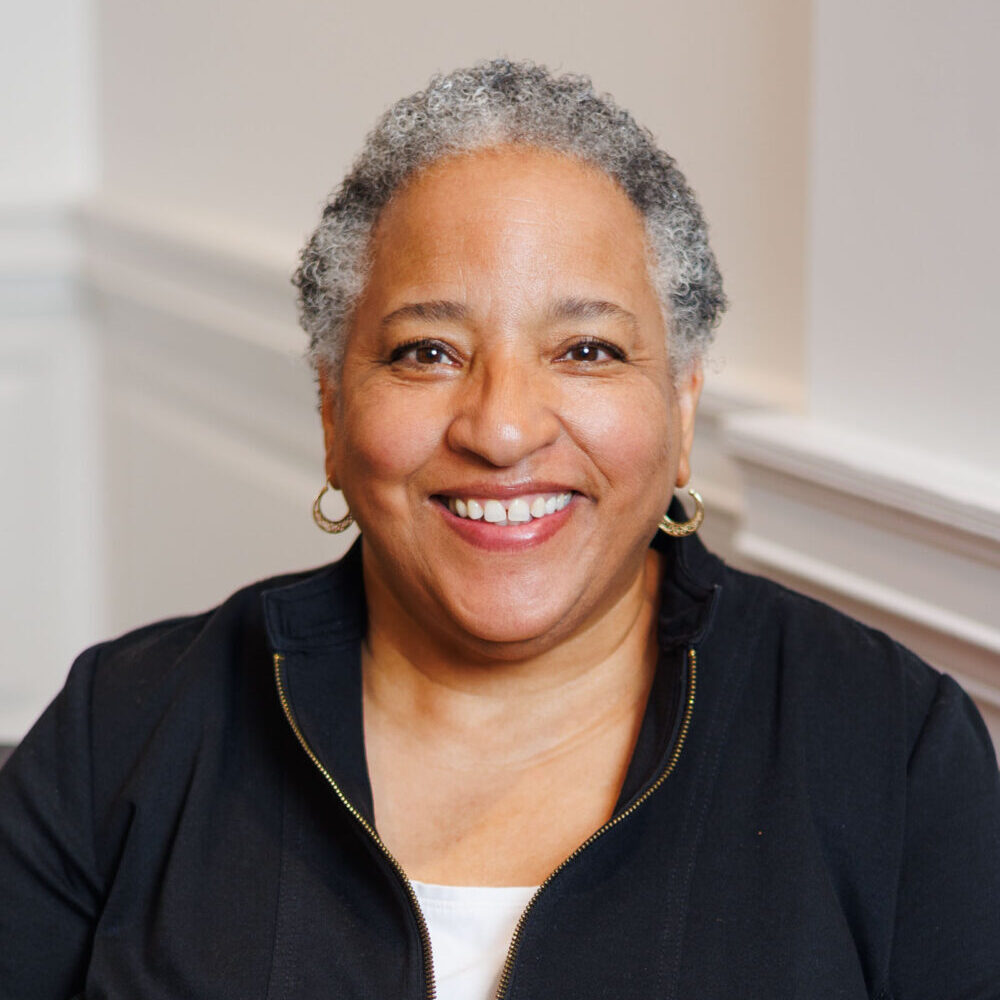An image of Keisha Blain weaing a textured white jacket and a blue-green shirt. She wears twists just past her shoulders and black glasses as she smiles for the camera.