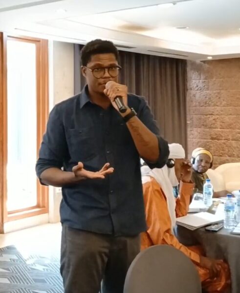 Dr. Leonard Mukosi, wearing glasses, a blue button-down shirt and gray-brown pants, stands, speaking into a microphone, as other convening members listen.
