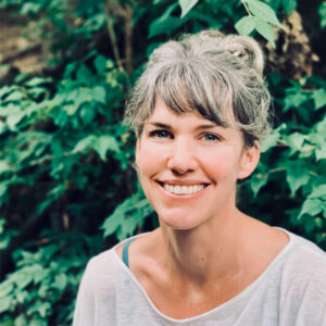 An image of Keisha Blain weaing a textured white jacket and a blue-green shirt. She wears twists just past her shoulders and black glasses as she smiles for the camera.