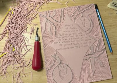 Image of linocut rubber block in process. Besides the block are a pencil, eraser, linocutting tool, and rubber scraps.
