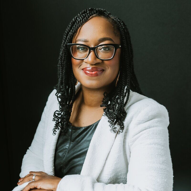 An image of Keisha Blain weaing a textured white jacket and a blue-green shirt. She wears twists just past her shoulders and black glasses as she smiles for the camera.